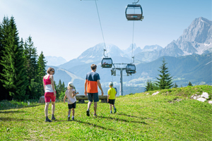 Vierköpfige Familie wandert auf Wiese unter einer Gondelbahn mit Bergblick im Sommer