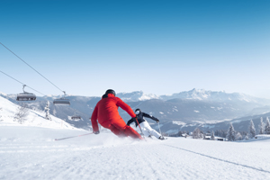 Zwei Skifahrer carven bei Sonne auf breiter, präparierter Skipiste mit Bergblick